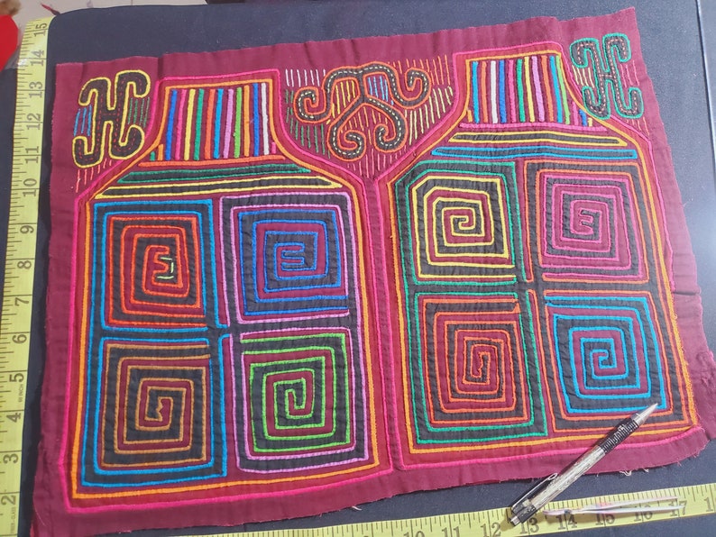 Puede incluir: Un tejido mola colorido con fondo rojo y dise&ntilde;os geom&eacute;tricos intrincados en tonos de azul, verde, amarillo, naranja y rosa. La mola presenta dos patrones sim&eacute;tricos de cuadrados conc&eacute;ntricos y espirales.