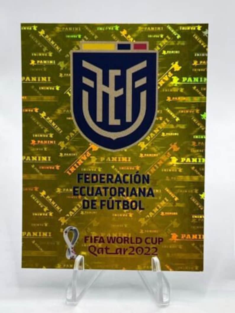 Puede incluir: Una pegatina Panini con el escudo de la Federaci&oacute;n Ecuatoriana de F&uacute;tbol en un escudo azul marino con una parte superior amarilla y roja. La pegatina tiene un fondo hologr&aacute;fico dorado con el texto "PANINI". Tambi&eacute;n es visible el logotipo de la Copa Mundial de la FIFA Qatar 2022.