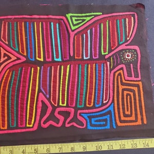 Puede incluir: Una mola colorida, un arte textil tradicional de la regi&oacute;n de Guna Yala en Panam&aacute;, que presenta un dise&ntilde;o geom&eacute;trico de un p&aacute;jaro con costuras vibrantes de rojo, naranja, amarillo, verde, azul y morado sobre un fondo de tela negro.
