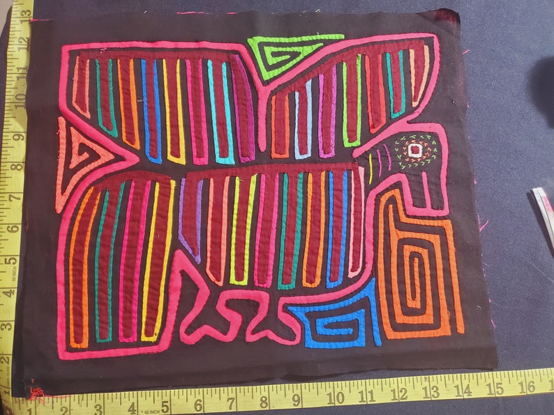 Puede incluir: Una mola colorida, un arte textil tradicional de la regi&oacute;n de Guna Yala en Panam&aacute;, que presenta un dise&ntilde;o geom&eacute;trico de un p&aacute;jaro con costuras vibrantes de rojo, naranja, amarillo, verde, azul y morado sobre un fondo de tela negro.