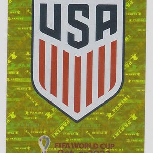 Puede incluir: Una pegatina Panini con el logotipo del equipo nacional de EE. UU. El emblema en forma de escudo tiene un borde blanco, las letras "USA" en azul marino y rayas rojas y blancas. El fondo es dorado con texto "Panini" y el logotipo de la Copa Mundial de la FIFA Qatar 2022.