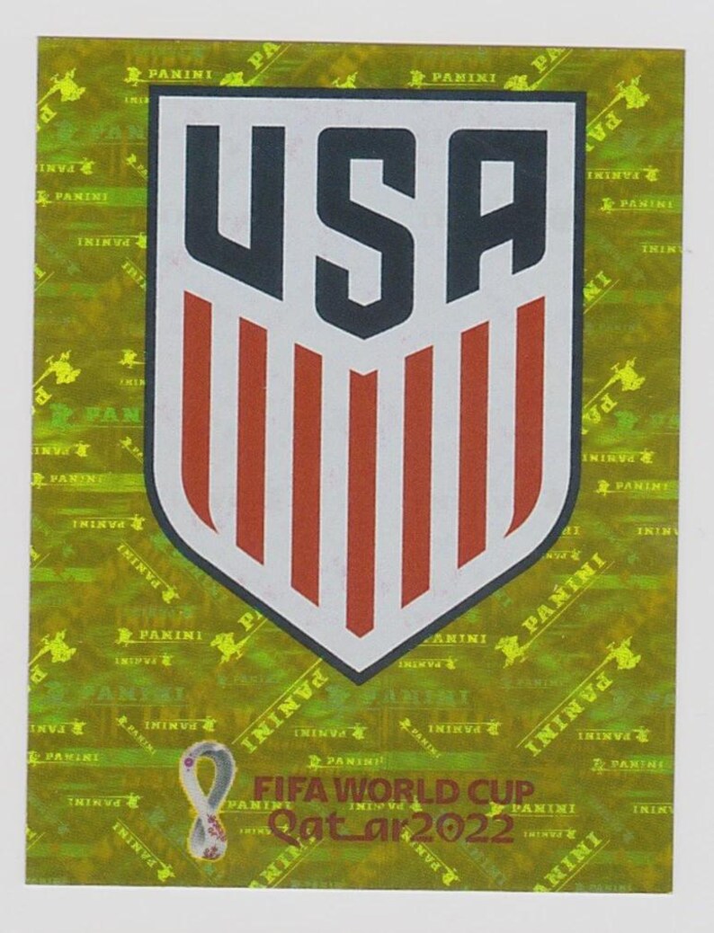 Puede incluir: Una pegatina Panini con el logotipo del equipo nacional de EE. UU. El emblema en forma de escudo tiene un borde blanco, las letras "USA" en azul marino y rayas rojas y blancas. El fondo es dorado con texto "Panini" y el logotipo de la Copa Mundial de la FIFA Qatar 2022.
