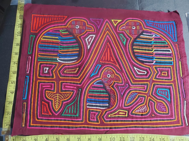 Puede incluir: Una obra de arte textil bordada de colores que representa tres p&aacute;jaros con dise&ntilde;os intrincados. Los p&aacute;jaros est&aacute;n delineados en negro y rellenos de colores vibrantes como rojo, azul, verde y amarillo. El fondo es de color burdeos oscuro.