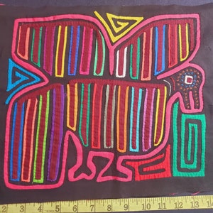 Puede incluir: Una mola colorida, un arte textil tradicional de la regi&oacute;n de Guna Yala en Panam&aacute;, que representa un p&aacute;jaro estilizado con un pico rojo y ojos negros. La mola est&aacute; hecha de capas de tela cosidas juntas y presenta una variedad de colores brillantes, incluyendo rojo, rosa, azul, verde, amarillo y morado.