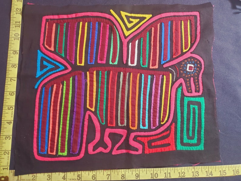 Puede incluir: Una mola colorida, un arte textil tradicional de la regi&oacute;n de Guna Yala en Panam&aacute;, que representa un p&aacute;jaro estilizado con un pico rojo y ojos negros. La mola est&aacute; hecha de capas de tela cosidas juntas y presenta una variedad de colores brillantes, incluyendo rojo, rosa, azul, verde, amarillo y morado.