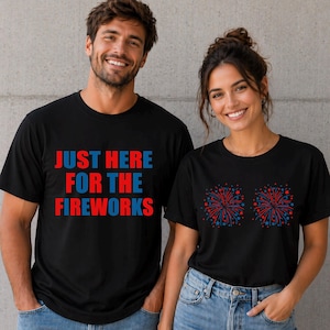 May include: Black t-shirts with patriotic designs. One shirt has the text "JUST HERE FOR THE FIREWORKS" in red and blue. The other shirt features two fireworks displays in red and blue. Both shirts are worn by smiling people.