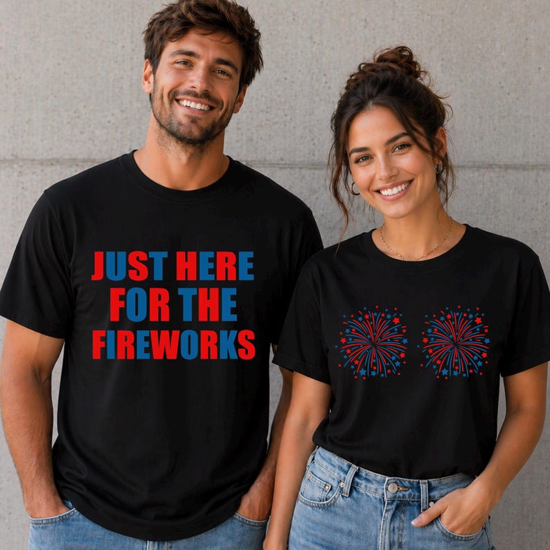 May include: Black t-shirts with patriotic designs. One shirt has the text "JUST HERE FOR THE FIREWORKS" in red and blue. The other shirt features two fireworks displays in red and blue. Both shirts are worn by smiling people.