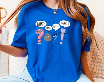May include: Royal blue t-shirt featuring a whimsical design of animated punctuation marks. The design includes a pink question mark, a gray rock, a blue comma, and a purple exclamation point, each with expressive faces and speech bubbles that say "WHAT?", "STOP.", "WAIT.", and "AGREE!"
