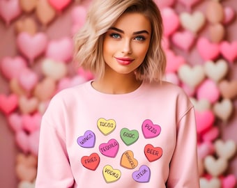 May include: A light pink sweatshirt with colorful heart-shaped graphics. Each heart features a food or drink item, including tacos, pizza, beer, and vodka. The background is a wall of pink and white heart-shaped balloons.
