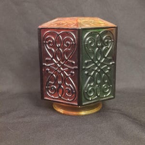 May include: A multi-colored, hexagonal glass perfume bottle with a gold base. The bottle features red, green, and orange panels with an ornate, swirling design. The bottle is likely vintage and decorative.