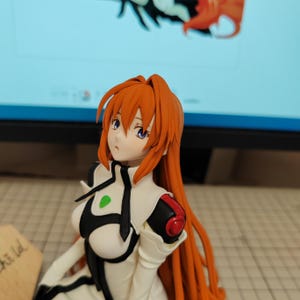 May include: A close-up of a red-haired anime figurine wearing a white and black outfit with a green accent. The figurine has blue eyes and is looking to the right.
