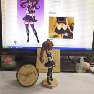May include: A brown haired anime figurine wearing a black and white sailor outfit with a purple bow. The figurine is standing on a wooden base. The figurine is in front of a computer screen showing a drawing of a similar anime character.