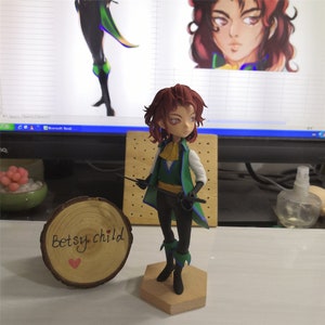 May include: A hand-painted figurine of a young person with brown hair, wearing a green and blue outfit, holding a sword. The figurine is standing on a hexagonal wooden base. The base has the words "Betsy child" written on it.