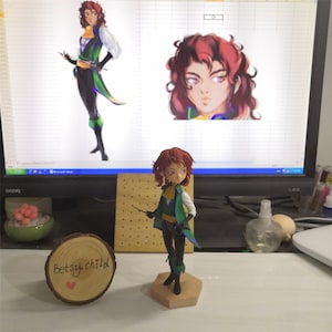 May include: A hand-painted figurine of a character with red hair, wearing a green and black outfit, holding a sword. The figurine is standing on a wooden base. The image also includes a computer screen with a digital illustration of the same character.