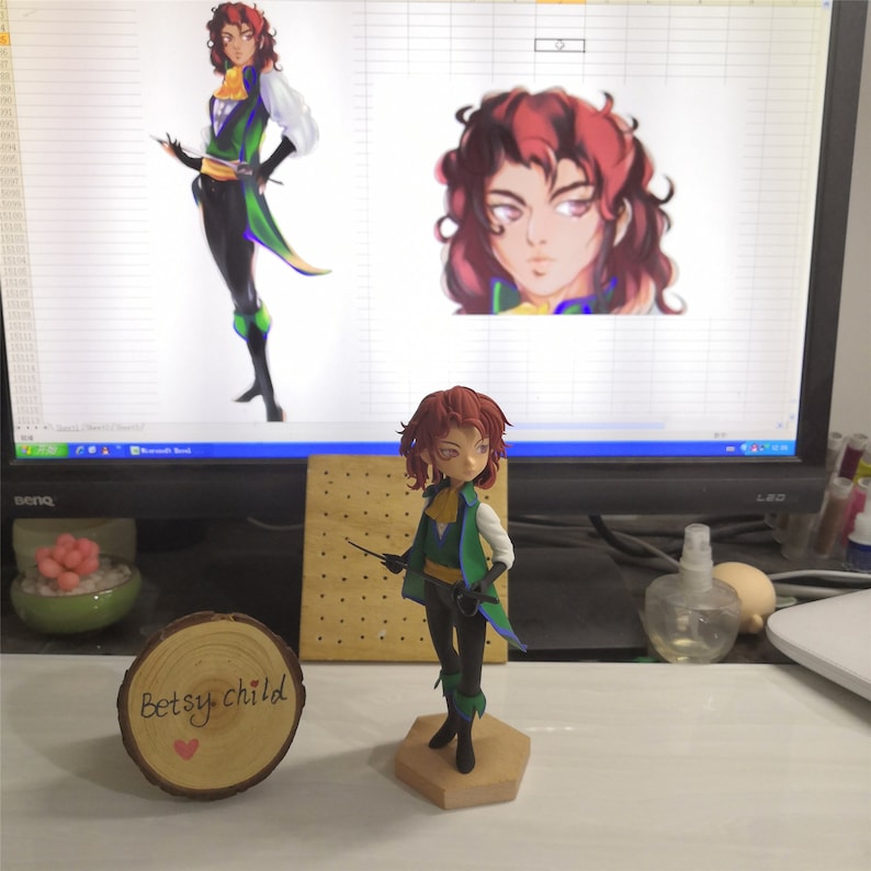 May include: A hand-painted figurine of a character with red hair, wearing a green and black outfit, holding a sword. The figurine is standing on a wooden base. The image also includes a computer screen with a digital illustration of the same character.