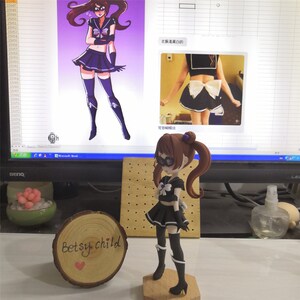 May include: A black and white figurine of a girl with brown hair and a black and white sailor outfit. The figurine is standing on a wooden base. The figurine is in front of a computer screen with a drawing of a girl in a similar outfit. The text on the screen says "衣服是黑白的" and "背部期结".
