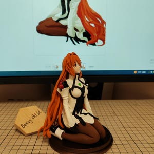 May include: A hand-painted figurine of a female anime character with long red hair, wearing a white and black outfit with a green gem on her chest. The figurine is kneeling on a wooden base. The text "Bessy child" is written on a wooden sign in the foreground.