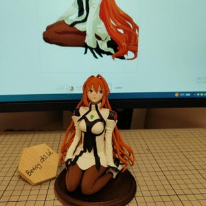 May include: A red-haired anime figurine of a woman wearing a white and black outfit with a green gem on her chest. She is kneeling on a wooden base. The figurine is in front of a computer monitor.