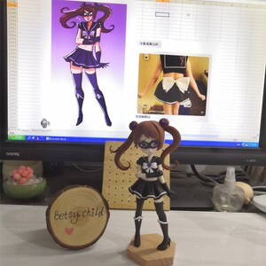 May include: A handmade doll figurine of a girl with brown hair, wearing a black and purple sailor outfit with a white bow. The doll is standing on a wooden base. The doll is next to a computer screen showing a drawing of a similar character.