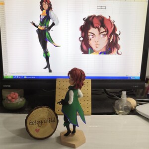May include: A green and blue figurine of a person with red hair. The figurine is standing on a wooden base. The figurine is holding a small gun in its right hand. The figurine is in front of a computer monitor. The monitor is displaying a digital drawing of a person with red hair. The drawing is on a grid background. The text "Betsy child" is written on a wooden circle.