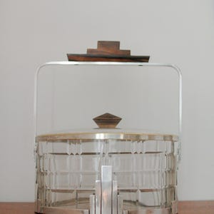 Puede incluir: Una cubitera de cristal transparente con base y asa de metal plateado. La tapa está rematada con un asa de madera geométrica. La cubitera tiene un patrón rectangular y se apoya sobre una superficie de madera. Un artículo decorativo.