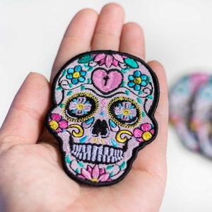 May include: A pink and purple embroidered sugar skull patch with colorful flowers and a heart. The patch is held in a hand.