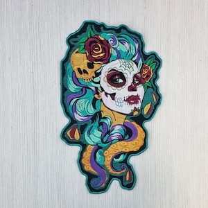 Op de afbeelding: Gestikt patch met een kleurrijke Día de Muertos suiker schedel met bloemen en een octopus. De schedel heeft een wit gezicht met zwarte ogen, een rode mond en een zwart-wit spinnenwebontwerp. De bloemen zijn rood, geel en groen. De octopus is paars en geel.