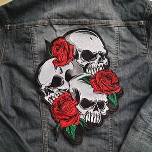May include: A black embroidered patch featuring three white skulls with black eyes and teeth, surrounded by red roses with green leaves.