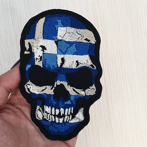 May include: A blue and white skull patch featuring the Greek flag design. The skull is embroidered with white and blue thread on a black fabric background.