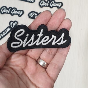 May include: Black embroidered patch with white lettering that reads "Sisters".