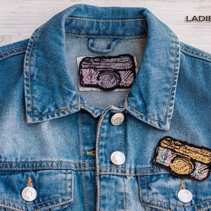 Op de afbeelding: Een blauwe denimvest met twee geborduurde camera-patches. De bovenste patch is roze en de onderste patch is goud.