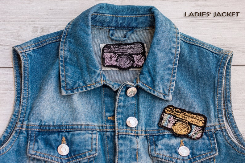 Op de afbeelding: Een blauwe denimvest met twee geborduurde camera-patches. De bovenste patch is roze en de onderste patch is goud.