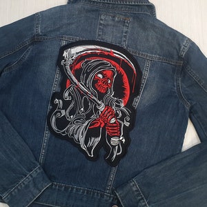 May include: A dark blue denim jacket with a large embroidered patch on the back. The patch depicts a red skull wielding a scythe, set against a red backdrop. The design is outlined in white.