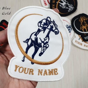 Könnte beinhalten: Weißer gestickter Aufnäher mit einem blauen Pferd und Reiter in einem goldenen Kreis. Der Text "YOUR NAME" ist in Gold unter dem Pferd gestickt.
