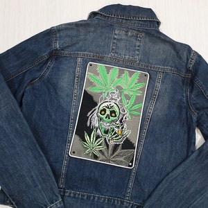 Può includere: Una giacca di jeans blu con una toppa ricamata verde e nera di uno scheletro che fuma uno spinello circondato da foglie di marijuana.