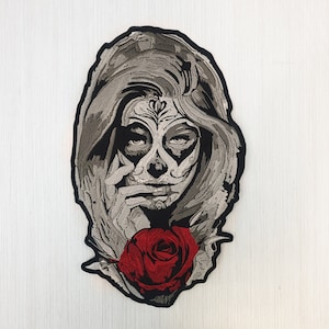 Könnte beinhalten: Gestickter Bügel-Patch mit einer Frau mit einem Zucker-Schädel-Make-up-Design und einer roten Rose. Die Frau hat langes, dunkles Haar und trägt ein schwarzes Kleid.
