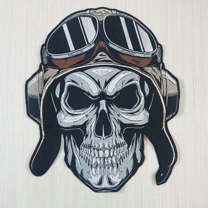 Könnte beinhalten: Ein gestickter Patch in Schwarzweiß, der einen Schädel zeigt, der einen braunen Lederfliegerhelm mit Brille trägt. Der Schädel hat einen bedrohlichen Ausdruck mit weit offenem Mund und scharfen Zähnen.