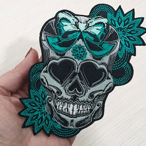 Könnte beinhalten: Ein gestickter Bügel-Patch mit einem Totenkopf mit einem türkisfarbenen Schmetterling und einem floralen Design. Der Schädel hat herzförmige Augen und ein zahniges Grinsen.