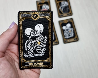 Puede incluir: Un parche rectangular bordado con fondo negro que muestra una pareja de esqueletos blancos besándose. El diseño incluye un corazón dorado en un esqueleto, un borde dorado, el número romano VI arriba y el texto THE LOVERS en la parte inferior.