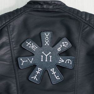 May include: Black leather jacket with a large, gray embroidered patch on the back. The patch features a starburst design with various symbols and runes.