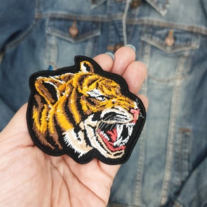 Puede incluir: Primer plano de un parche de hierro bordado que presenta la cabeza de un tigre con una expresión feroz. El tigre está representado en tonos de naranja, marrón, negro y blanco.