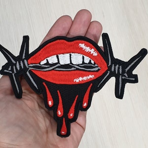 Puede incluir: Labios rojos con dientes blancos están atravesados por alambre de púas, con sangre roja goteando. El parche es negro con un contorno blanco.