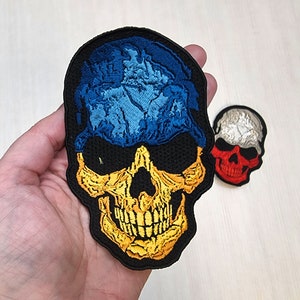 May include: A black skull embroidered patch with a blue and yellow design, resembling the Ukrainian flag. The skull is outlined in yellow and has a detailed design.