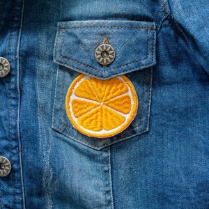 Può includere: Una giacca di jeans blu con una toppa ricamata a forma di fetta d'arancia gialla sulla tasca.