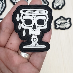 Op de afbeelding: Zwarte stoffen patch met een witte, geborduurde schedel in de vorm van een kelk. De schedel heeft een druppelend effect met witte druppels.