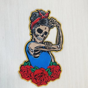 May include: A blue and yellow embroidered patch featuring a skeleton woman with a red bandana, flexing her bicep, and surrounded by red roses.
