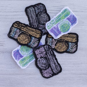 Op de afbeelding: Zes geborduurde strijkpatches in de vorm van vintage camera's. De patches zijn in verschillende kleuren, waaronder zwart, wit, goud, groen en paars.