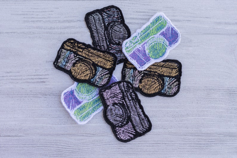 Op de afbeelding: Zes geborduurde strijkpatches in de vorm van vintage camera's. De patches zijn in verschillende kleuren, waaronder zwart, wit, goud, groen en paars.