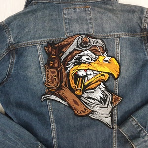 Puede incluir: Un parche bordado que representa un águila con un casco de aviador de cuero y gafas, sosteniendo un cigarro en su pico. El águila mira hacia la derecha. El parche es marrón, blanco y amarillo.