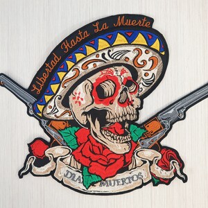 May include: A black embroidered patch with a skull wearing a colorful sombrero, two crossed pistols, a red rose, and a banner that reads "Libertad Hasta La Muerte Dia Muertos".